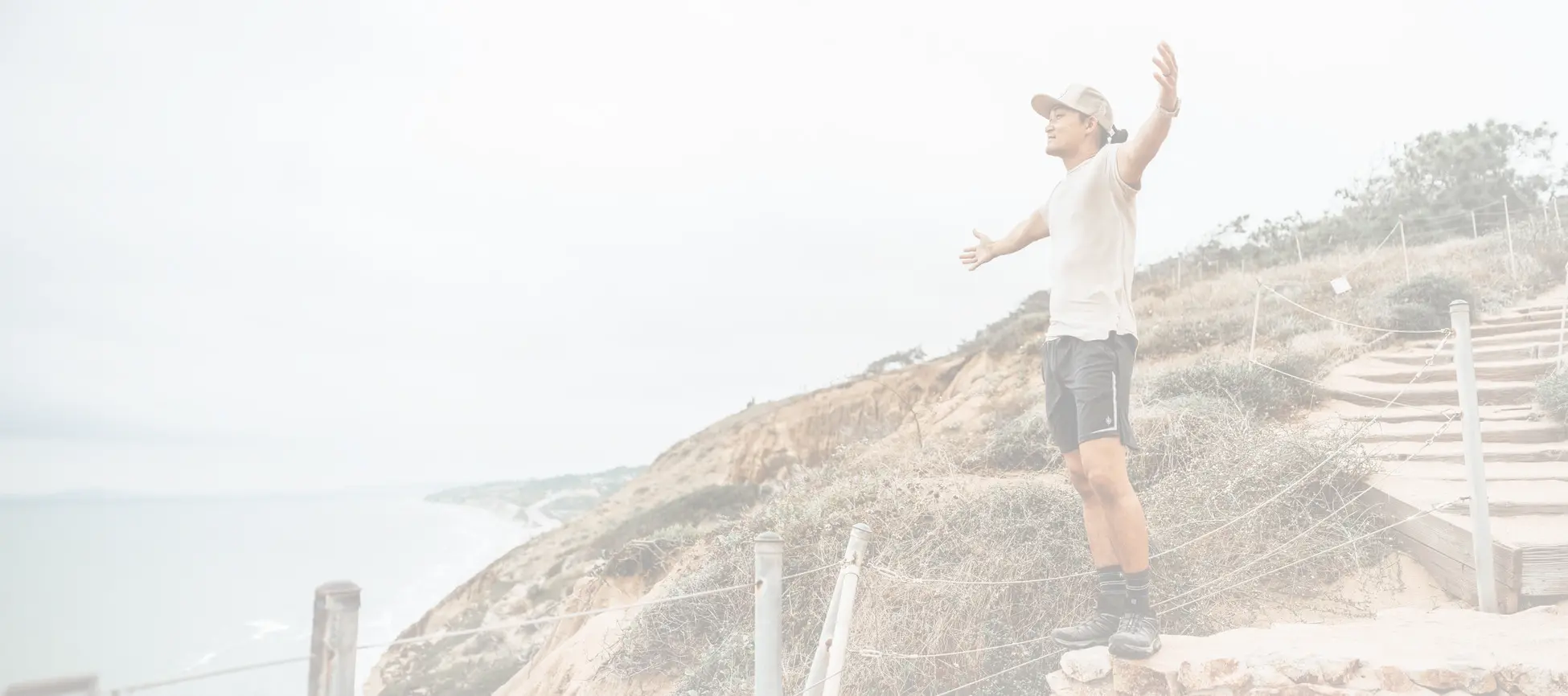 A man wearing a white t-shirt, black shorts, and a beige cap stands on a rocky coastal path with his arms outstretched, overlooking a beach and ocean under an overcast sky. Wearing Bamtech apparel.